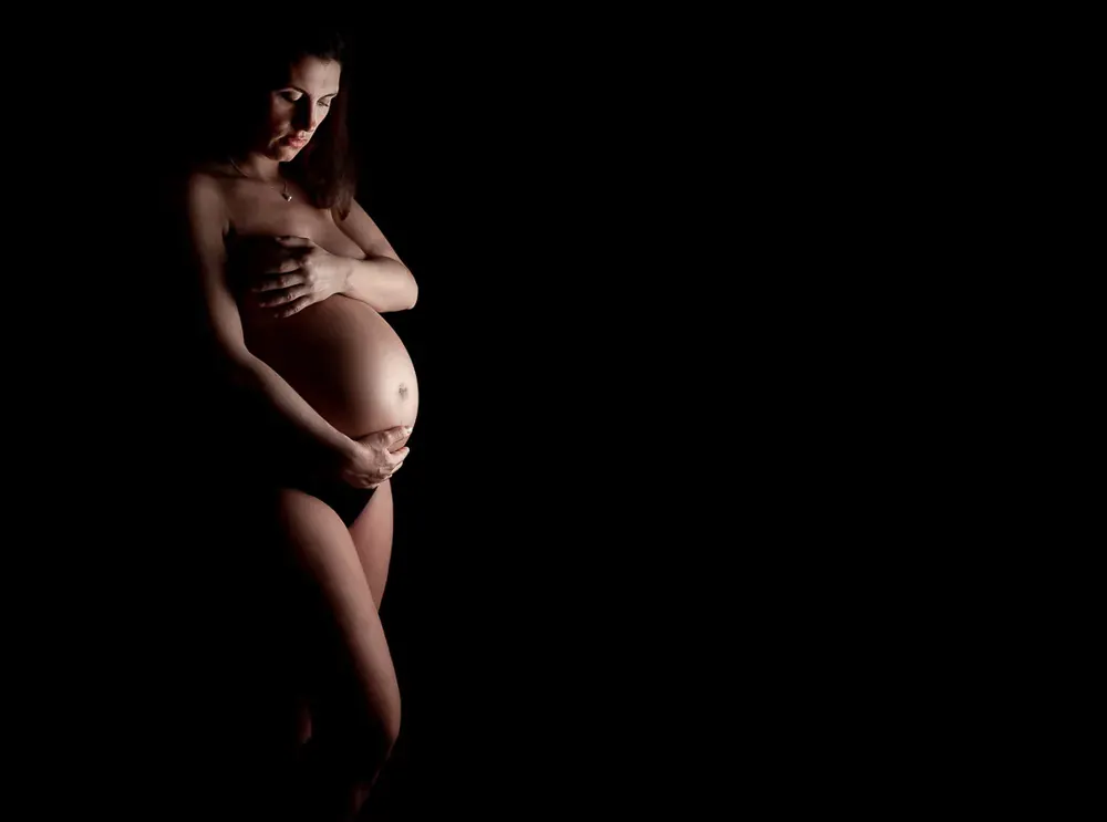 Donna in gravidanza a figura intera nella penombra, una mano sul petto e l'altra sulla pancia