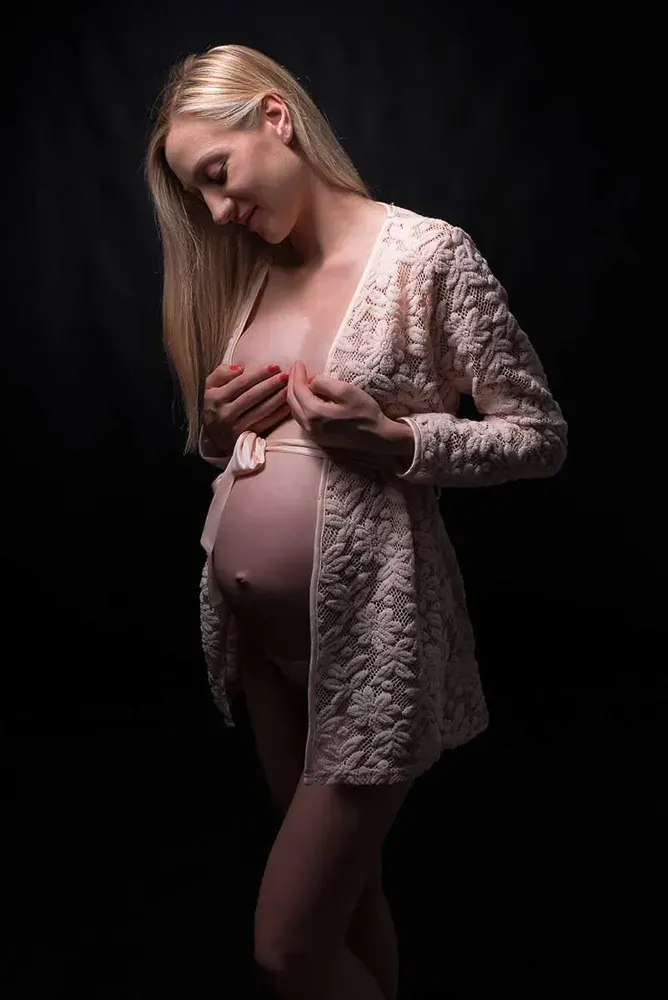 Donna bionda in gravidanza con una giacca di pizzo chiaro aperta sulla pancia, su fondo nero