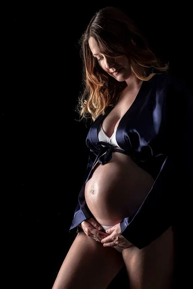 Donna in gravidanza con una giacca blu aperta, in intimo bianco, sorride guardando la pancia