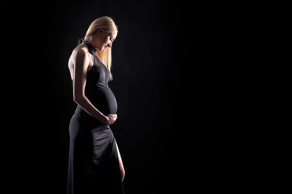 Donna bionda in gravidanza di profilo, in un lungo abito nero con spacco