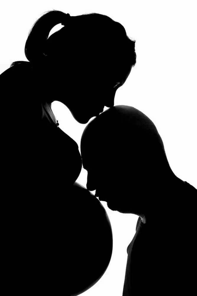Sagome nere su fondo bianco: una donna in gravidanza bacia sulla fronte l'uomo chino sulla sua pancia