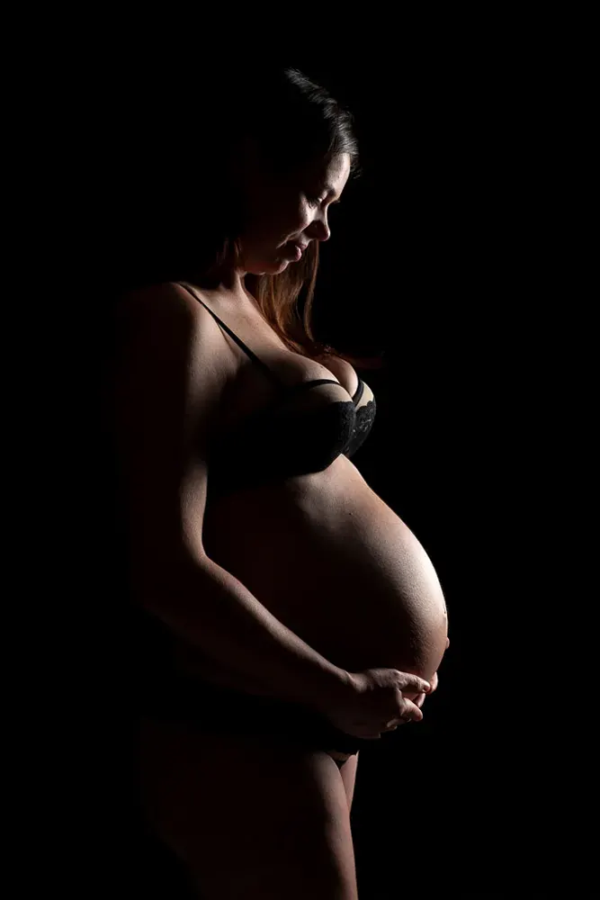 Donna in gravidanza in intimo nero, di profilo e in penombra, le mani sotto la pancia