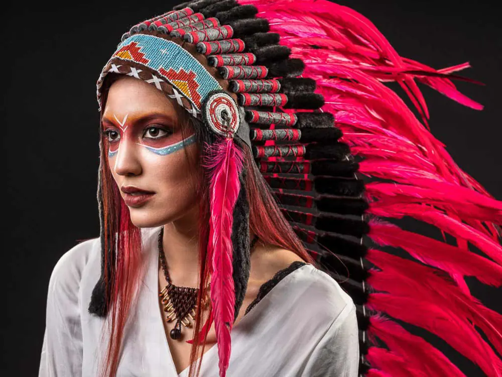 Donna con un grande copricapo di piume rosa e il viso dipinto a strisce colorate, su fondo scuro