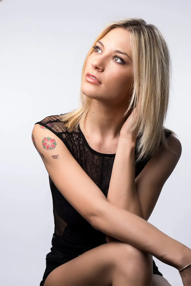 Ragazza bionda che guarda in alto, con un tatuaggio a fiore sulla spalla e un abito nero di pizzo