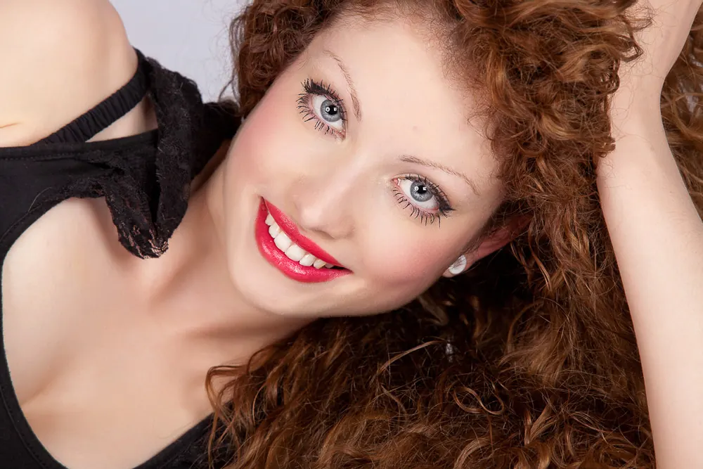 Primo piano di donna sdraiata coi capelli ricci rossi, gli occhi azzurri e un braccio alzato