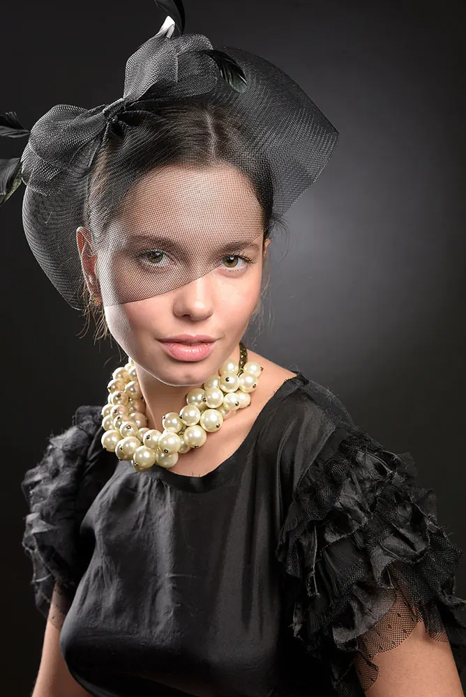 Donna con un cappellino nero col velo sugli occhi, una collana di grosse perle e una blusa nera a balze