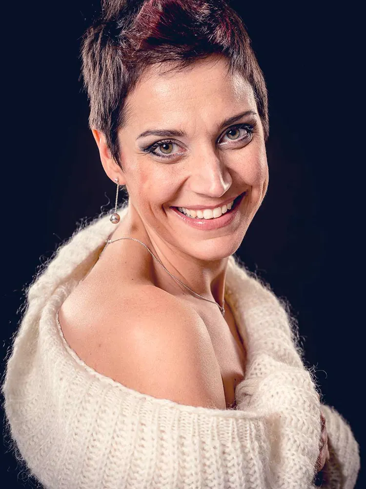 Donna coi capelli corti che ride, avvolta in un maglione bianco che le lascia scoperta una spalla