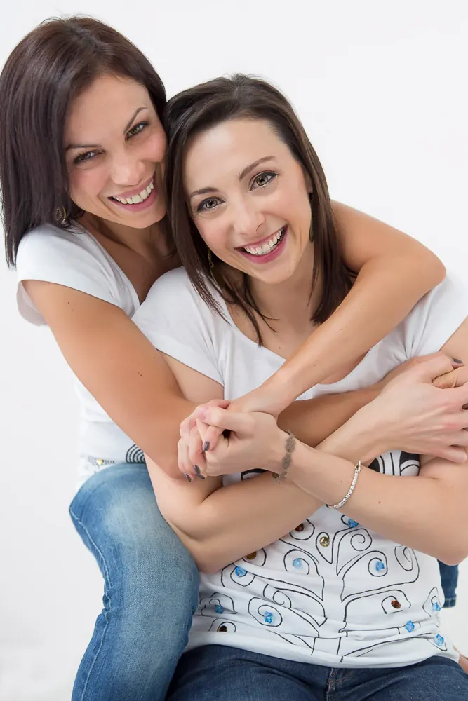 Due donne ridono abbracciate, una dietro l'altra, in magliette bianche e jeans