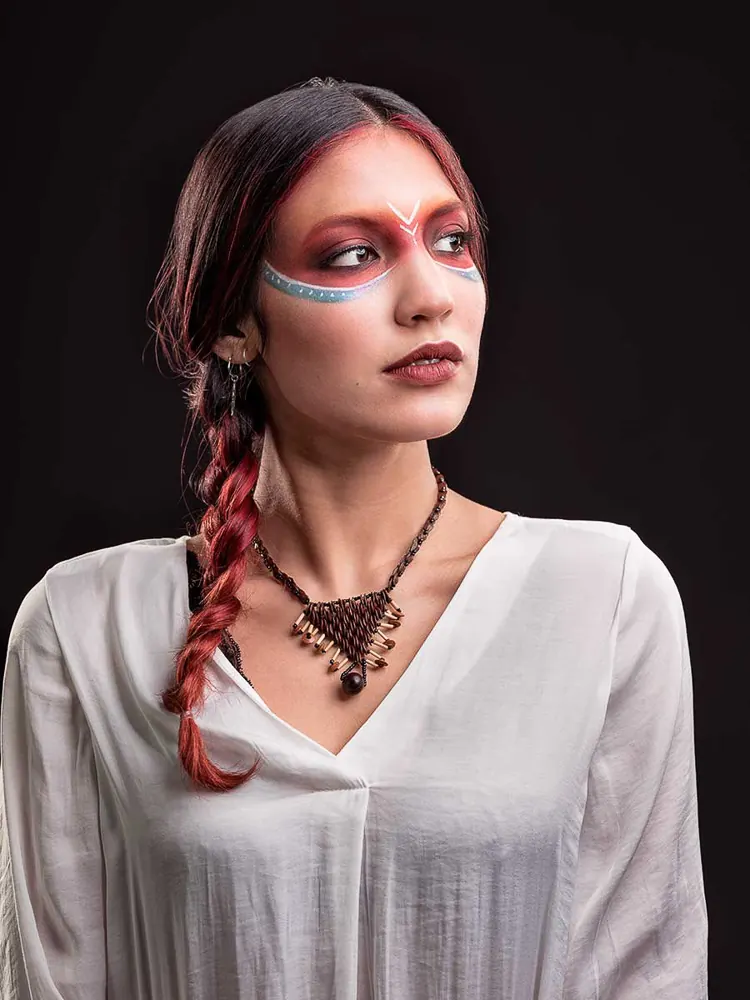 Donna che guarda di lato, col trucco colorato attorno agli occhi, una treccia rossa e una collana intrecciata