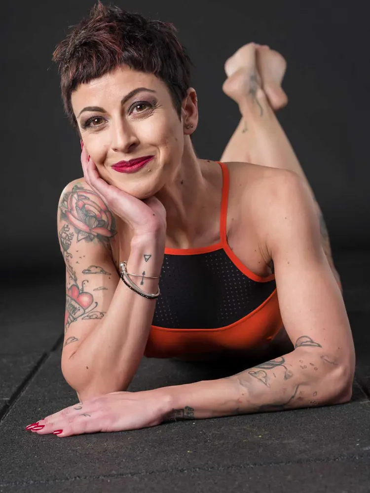 Donna coi capelli corti e tatuaggi sulle braccia, sdraiata sul ventre con il mento sulla mano, top arancione e nero