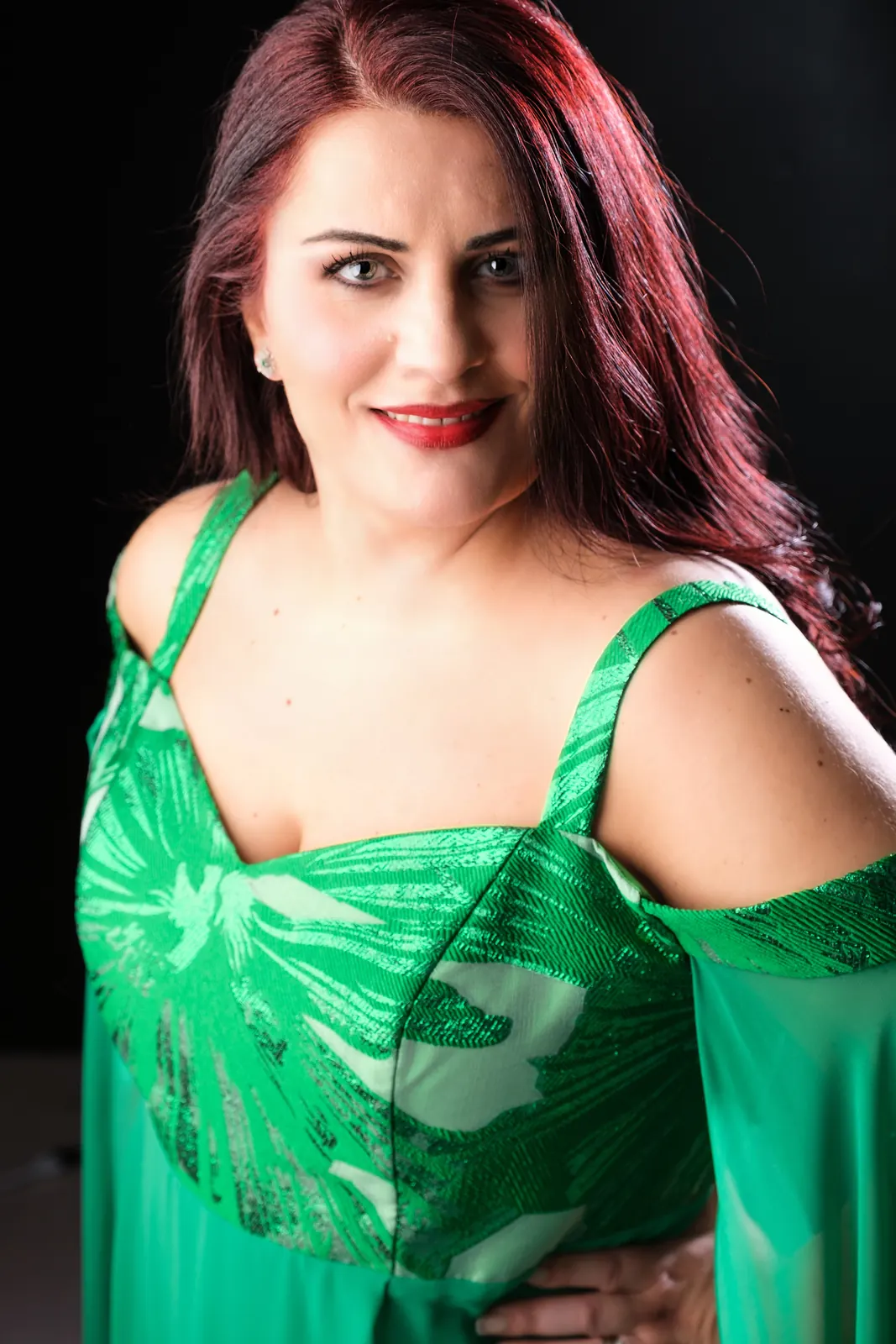 Ritratto di una cantante con abito verde a spalline e capelli bordeaux, sorride su fondo nero