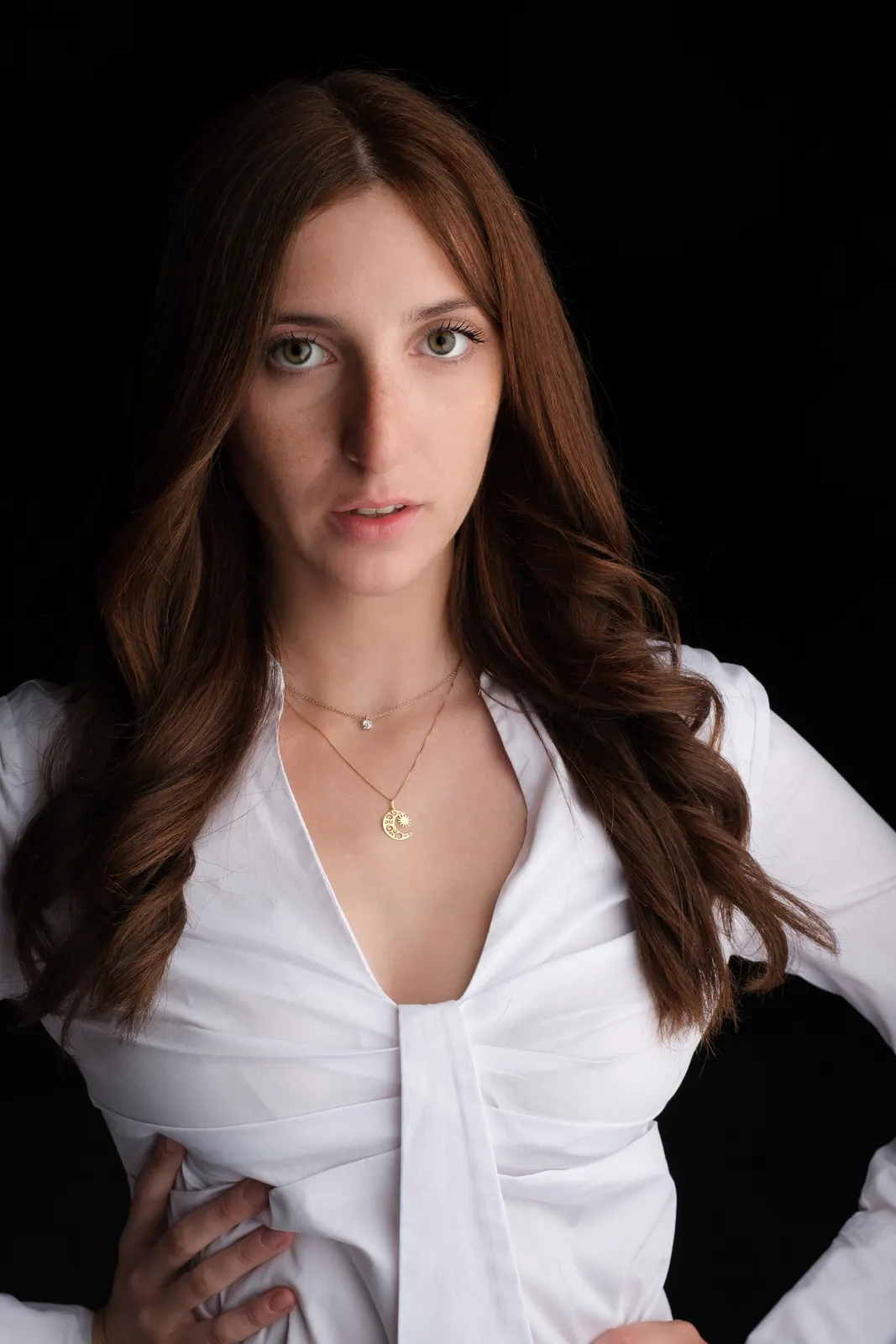Ragazza con camicia bianca scollata e ciondolo dorato, mani sui fianchi, su fondo nero