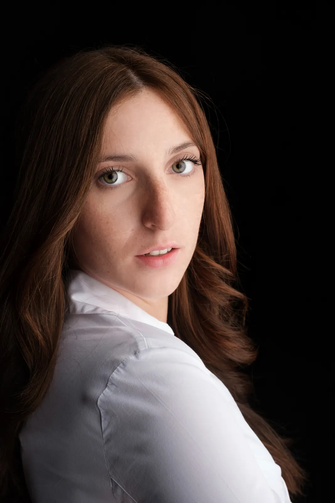 Ragazza in camicia bianca, voltata di spalle col viso verso l'obiettivo, luce laterale su fondo nero