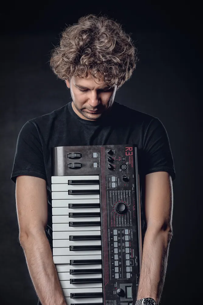 Musicista a testa china che regge una tastiera davanti al petto, su fondo scuro