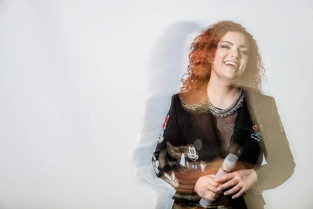 Cantante coi capelli rossi che ride tenendo il microfono, con la figura sdoppiata dal mosso su fondo bianco