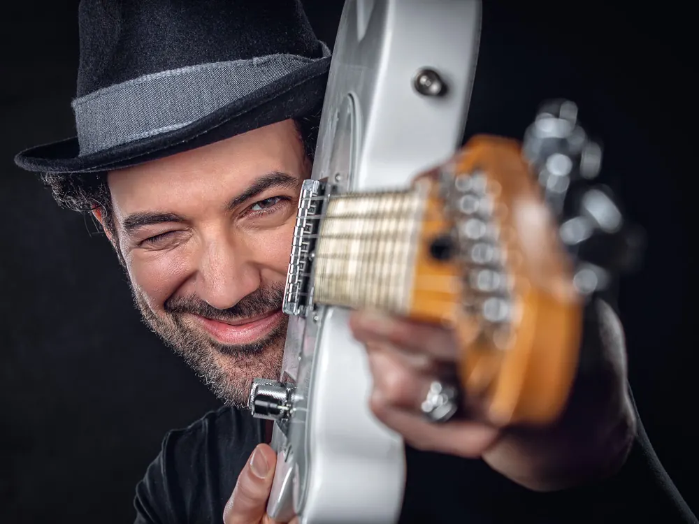 Musicista con il cappello che sbircia da dietro una chitarra bianca, sorridendo