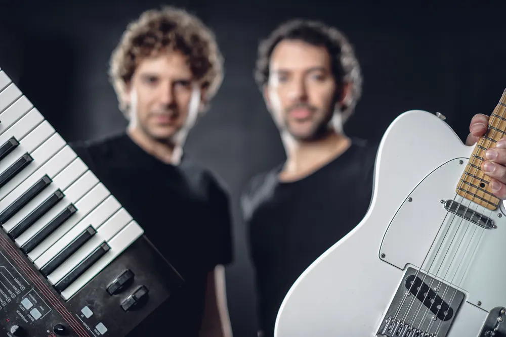 Due musicisti sfocati sullo sfondo, in primo piano una tastiera e una chitarra elettrica bianca