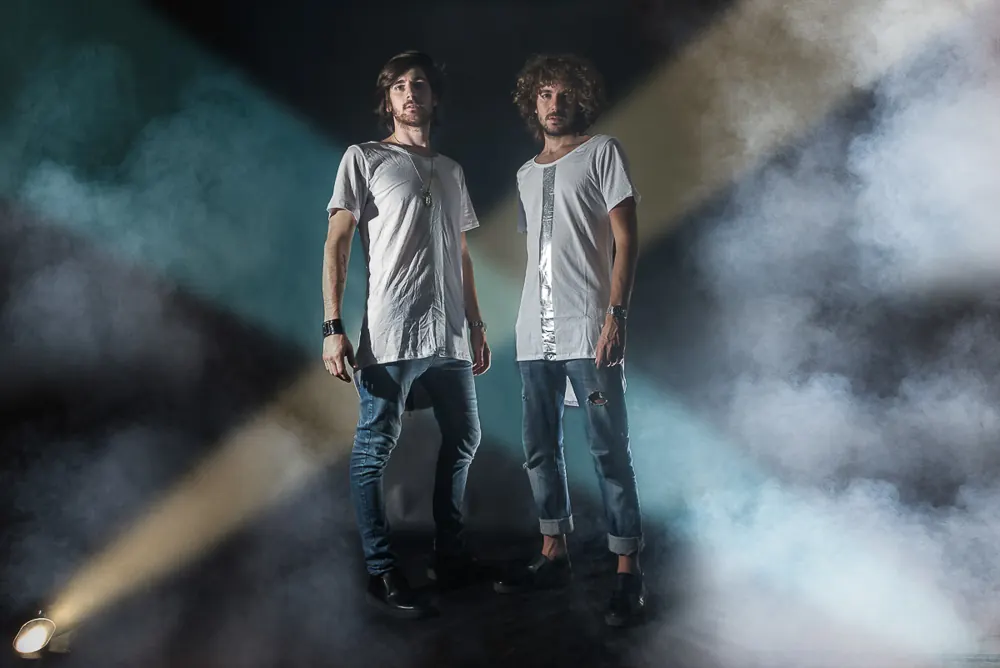 Due musicisti in piedi con magliette chiare, fra fumo e fasci di luce colorata