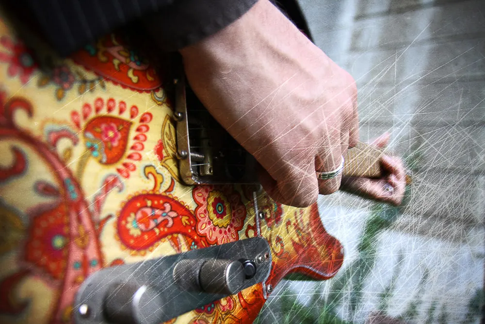 Dettaglio di una mano sulla chitarra elettrica a fantasia paisley, con graffi sovrapposti all'immagine