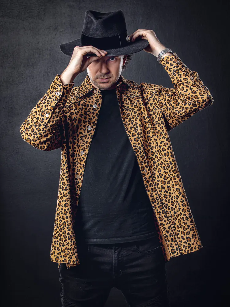 Uomo con cappello nero e camicia leopardata che si tiene la tesa con entrambe le mani