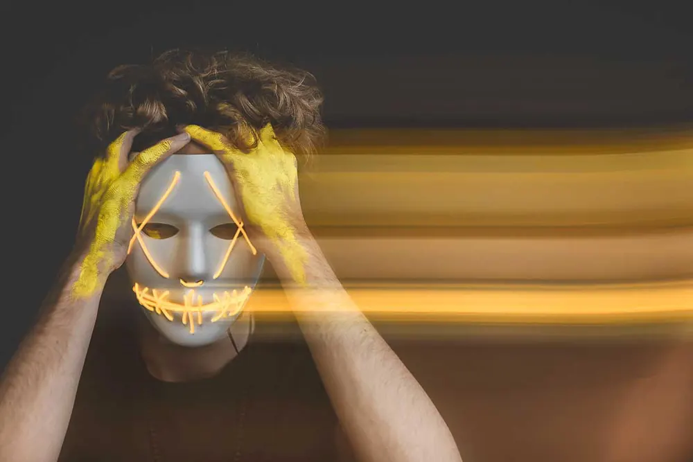 Uomo con una maschera bianca dai contorni luminosi e le mani dipinte di giallo sul volto, con scie di luce gialla