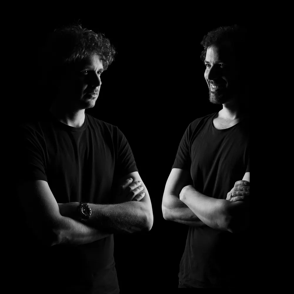 Due musicisti in bianco e nero con le braccia incrociate, uno di fronte all'altro, su fondo nero
