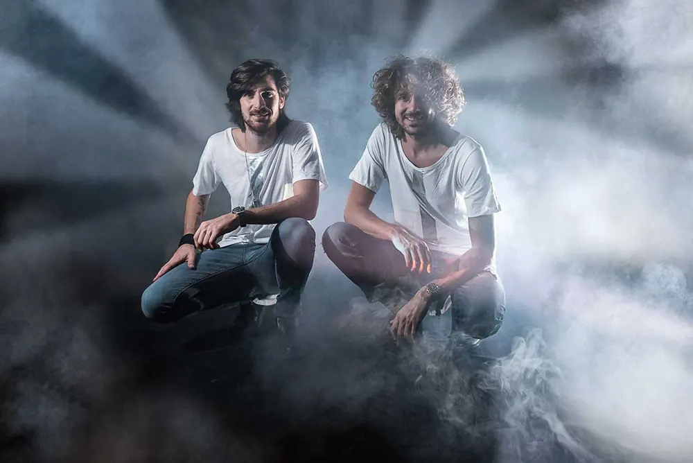 Due musicisti accovacciati in magliette bianche, immersi nel fumo e in fasci di luce