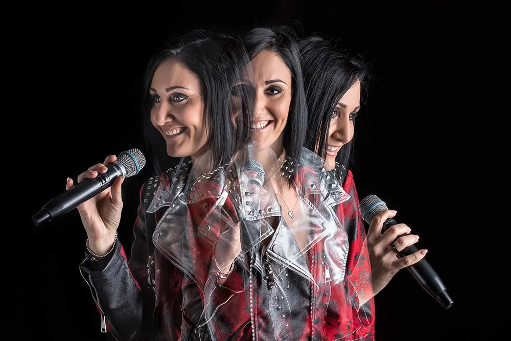 Cantante con giacca borchiata e due microfoni, la figura moltiplicata dal mosso su fondo nero