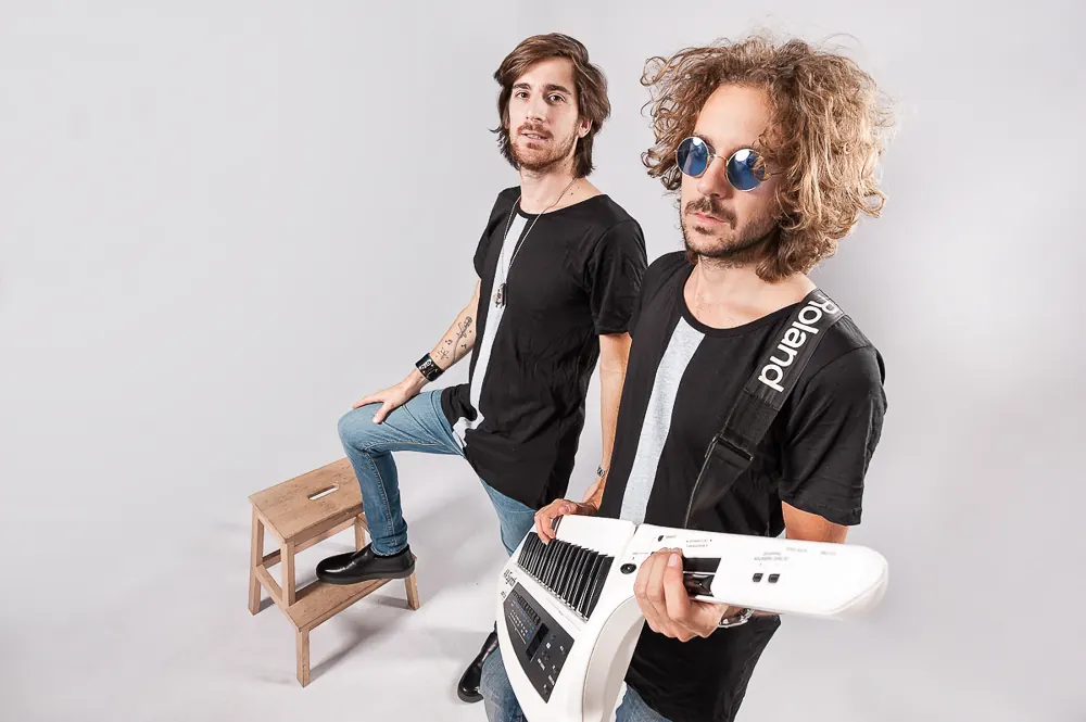 Musicista con tastiera bianca a tracolla e occhiali tondi blu, accanto a un altro con un piede su uno sgabello