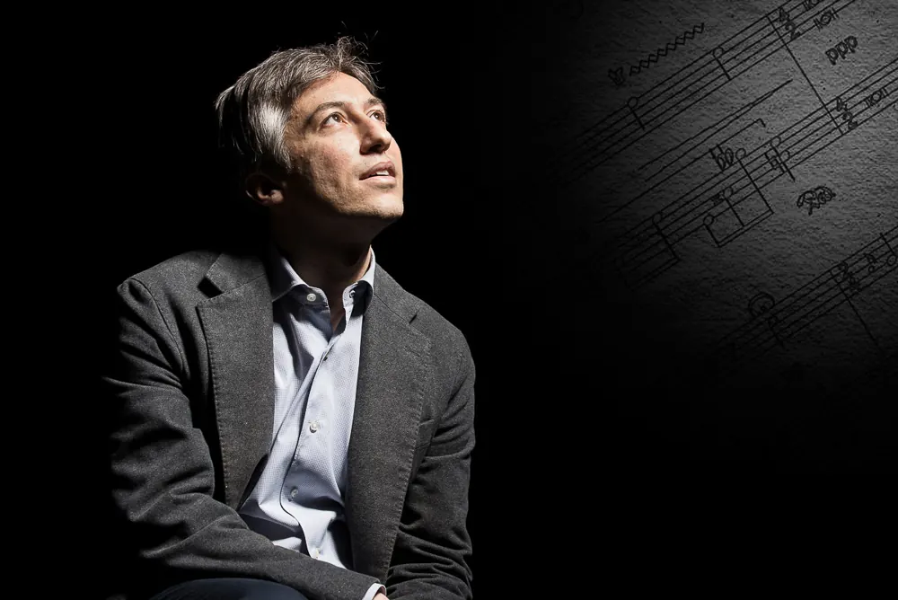 Uomo in giacca grigia che guarda in alto, con uno spartito musicale sul fondo a destra