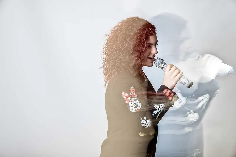Cantante coi ricci che canta nel microfono, la figura mossa e moltiplicata su fondo chiaro