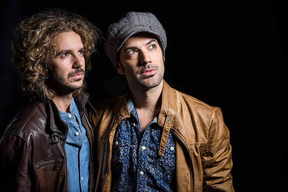 Musicista con coppola grigia e giacca di cuoio, al suo fianco uno coi ricci e il giubbotto scuro