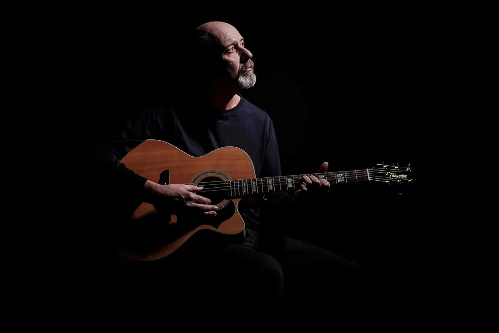 Chitarrista calvo con la barba grigia che suona una chitarra acustica, in penombra
