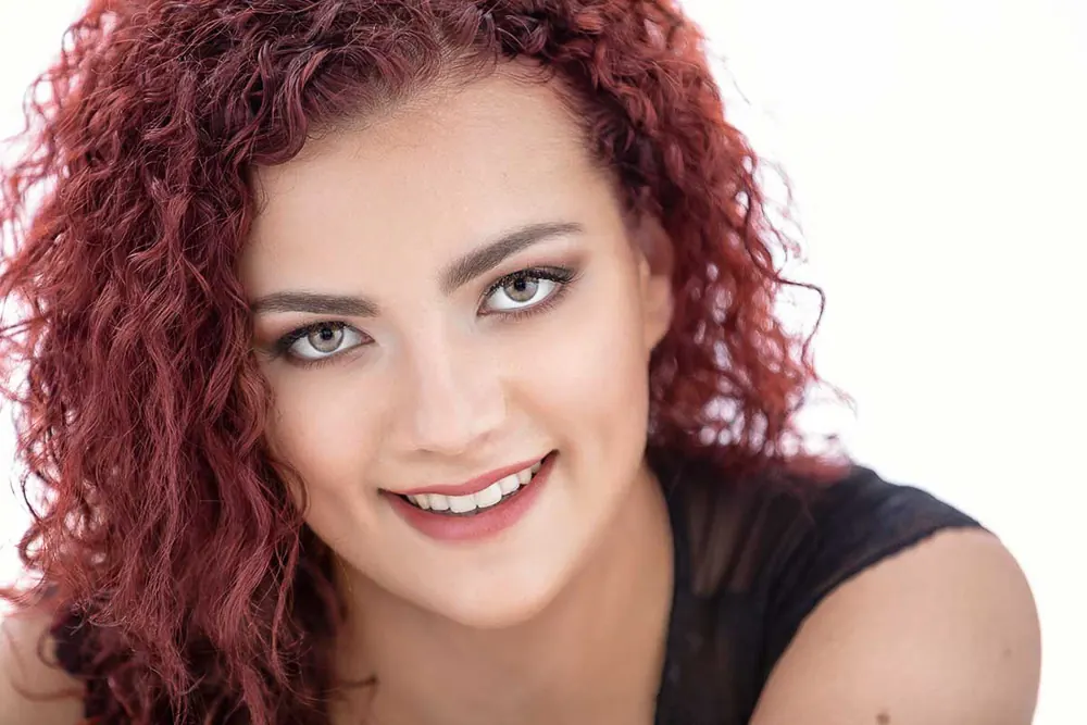 Primo piano di ragazza coi ricci rossi e occhi chiari che sorride, su fondo bianco
