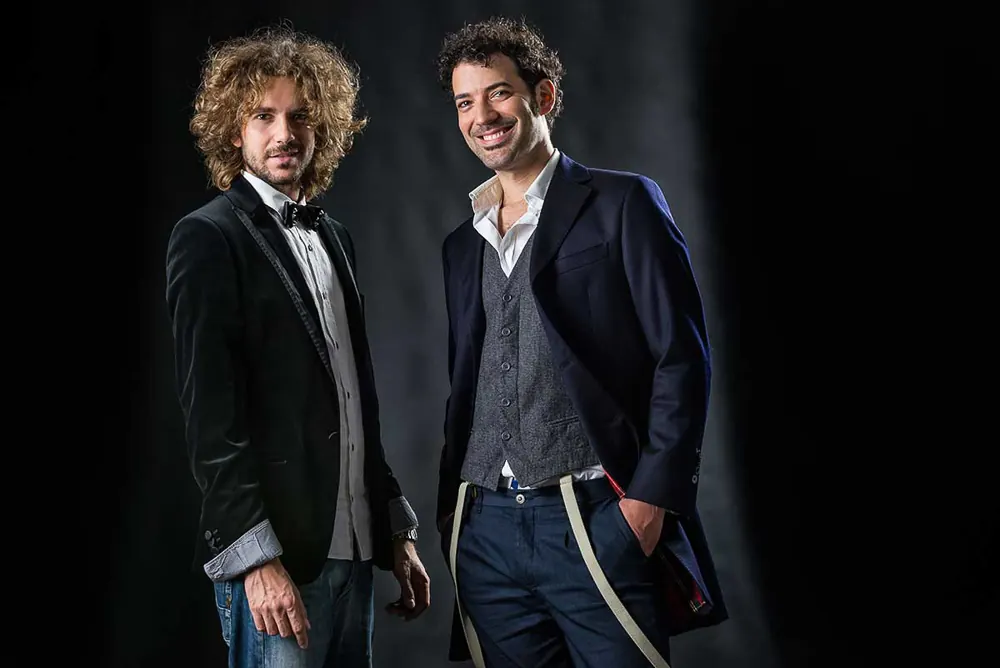 Due musicisti in giacca che sorridono, uno col papillon e l'altro con gilet grigio e bretelle