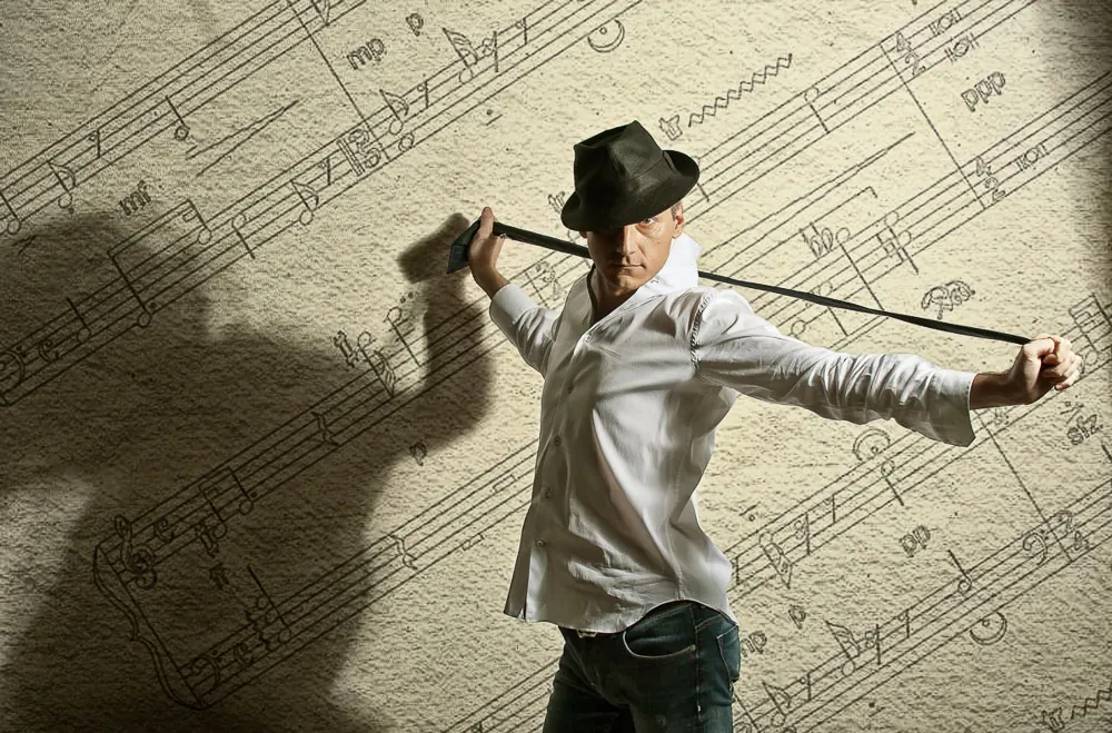Uomo con cappello e camicia bianca che tende una cravatta a braccia aperte, davanti a un muro di note musicali