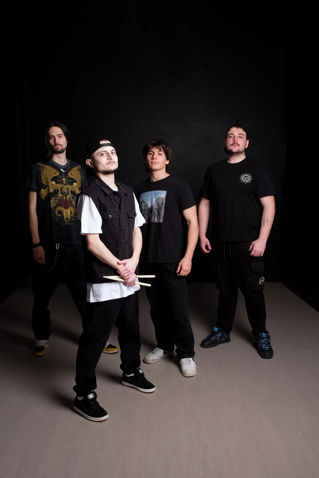Band di quattro ragazzi in piedi su fondo nero, il batterista davanti con le bacchette in mano